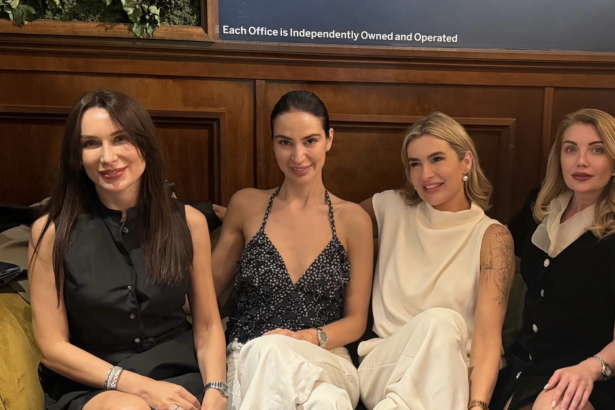 Four women sit closely on a bench in a wood-paneled setting, smiling at the camera, dressed in black and white outfits.