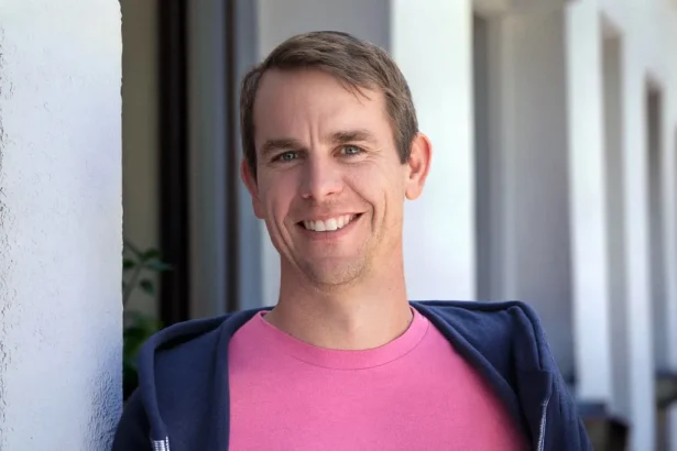 Smiling man in a pink shirt and navy jacket leaning against a white wall outdoors, facing the camera.