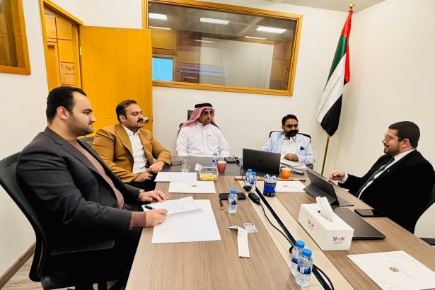 Five men in suits sit around a wooden conference table in a meeting room, with laptops, papers, water bottles, and snacks present; UAE flag in the corner.