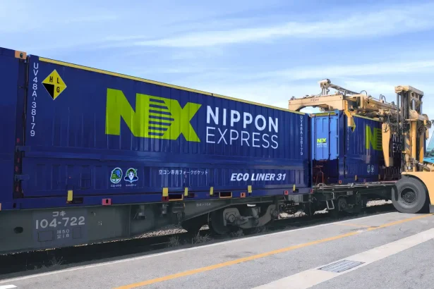Blue Nippon Express cargo container on a flatbed railcar being loaded by a yellow forklift/crane against a clear blue sky, showing the text 'Nippon Express' and 'NEX' logo in bright green.