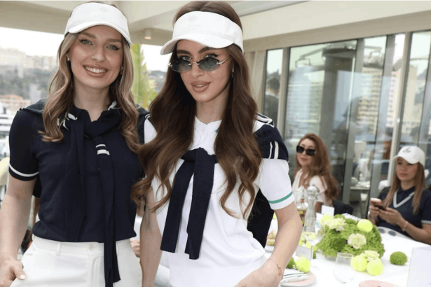 Two smiling women pose for a photo at a bright event, wearing navy and white outfits with white visors, tables and guests in the background.
