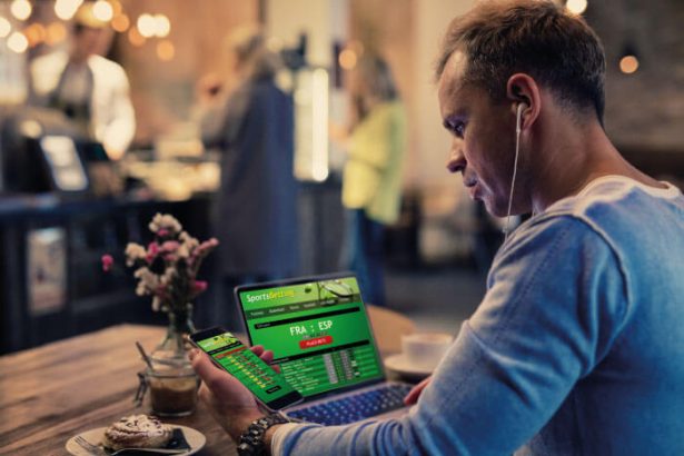 Man with earbuds using a tablet displaying a sports betting app at a cafe table.