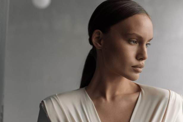 Close-up side profile of a woman with dark hair pulled back, wearing a light beige top, gazing to the right against a muted background.