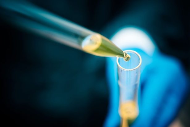 Gloved hand uses a pipette to dispense yellow liquid into a test tube.