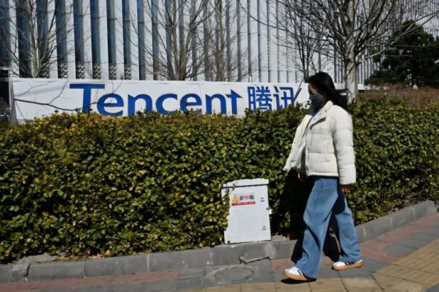Person wearing a white puffer jacket, blue jeans, and a face mask walks along a sidewalk in front of a Tencent sign and hedge.