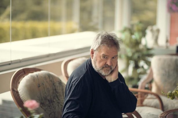 Older man with gray beard sitting in a sunlit lounge, resting his head on his hand by the window.
