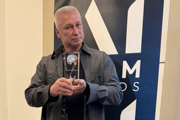 Man in a gray jacket holds a glass award with a brain image, standing before a blue and white banner.