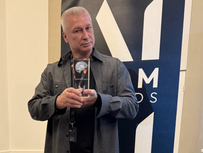 Man in a gray jacket holds a glass award with a brain image, standing before a blue and white banner.