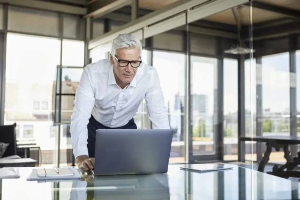Man in a white shirt and glasses leans over a silver laptop on a glass desk in a bright, modern office with large windows.