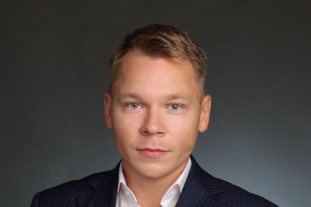 Professional headshot of a man in a dark pinstripe suit and white shirt, looking at the camera against a gray background.