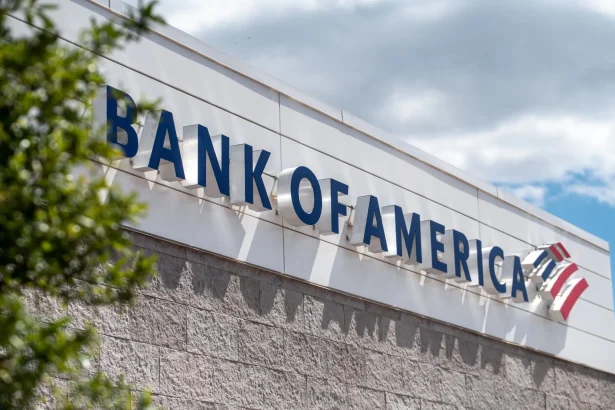 Side view of a Bank of America sign on a white building facade, blue letters visible with green tree foreground and a cloudy sky above.