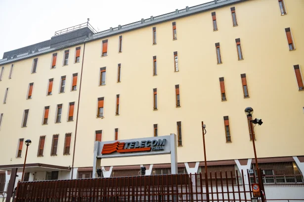Facade of a tall yellow building with narrow rectangular windows and a TELECOM ITALIA sign at the street level, in front of a brown fence.