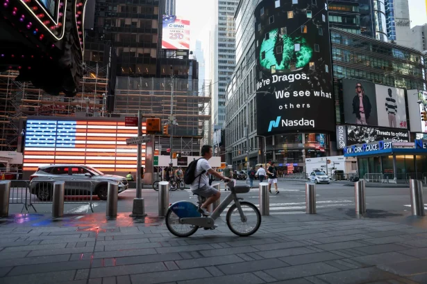 Busy Times Square street with a large Nasdaq billboard, cyclists and pedestrians amid tall buildings and an American flag display in the background.