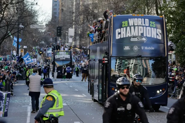 Blue double-decker parade bus with a 'World Champions' banner drives down a city street as crowds cheer from both sides and police officers stand in the foreground, confetti on the ground.