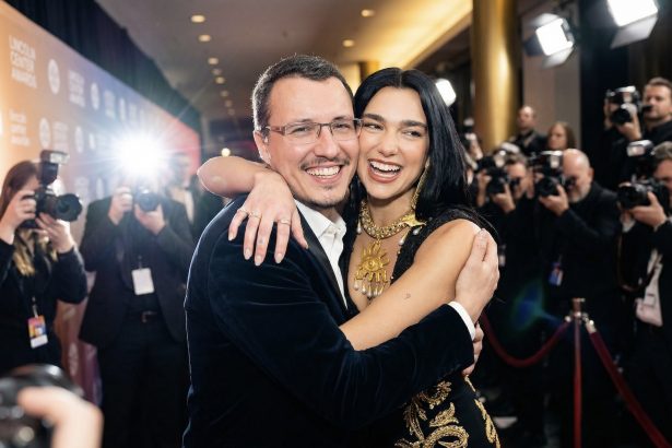 Man and woman hug and smile on a red carpet surrounded by photographers; glamorous formal attire and flashbulbs in the background.