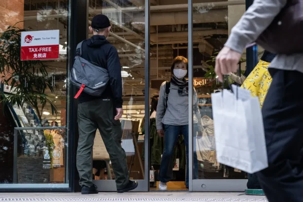 Shoppers enter a storefront through glass doors; a masked woman stands inside as a man with a backpack approaches the entrance.