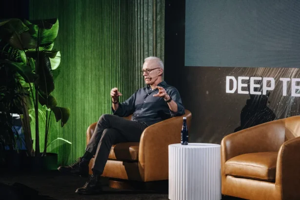 Speaker on stage: gray-haired man with glasses seated in a brown leather armchair, gesturing while talking; white ribbed table with a bottle nearby, green plants on the left, and a dark backdrop with partial text.