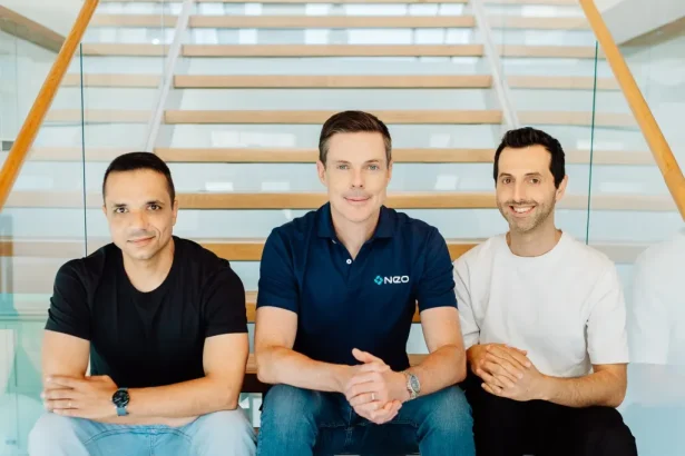 Three men sit on a wooden staircase in a modern office, smiling at the camera on a sunny day.