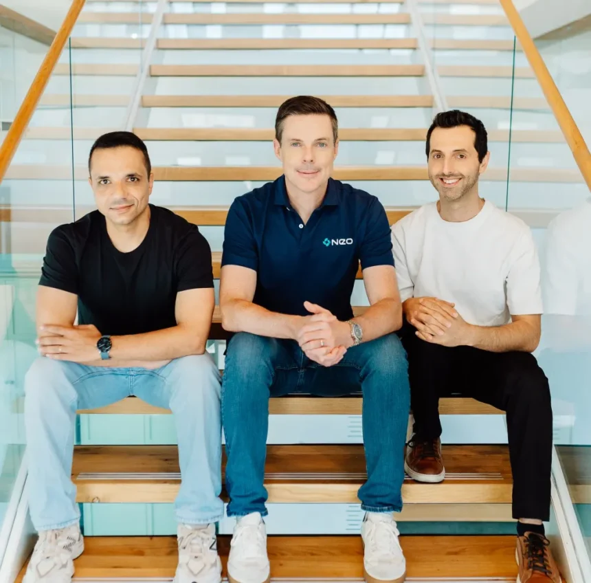 Three men sit on a wooden staircase in a modern office, smiling at the camera on a sunny day.