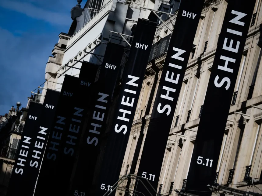 Tall black banners along a building spelling SHEIN with BHV logos and 5.11 at the bottom, against a blue sky.