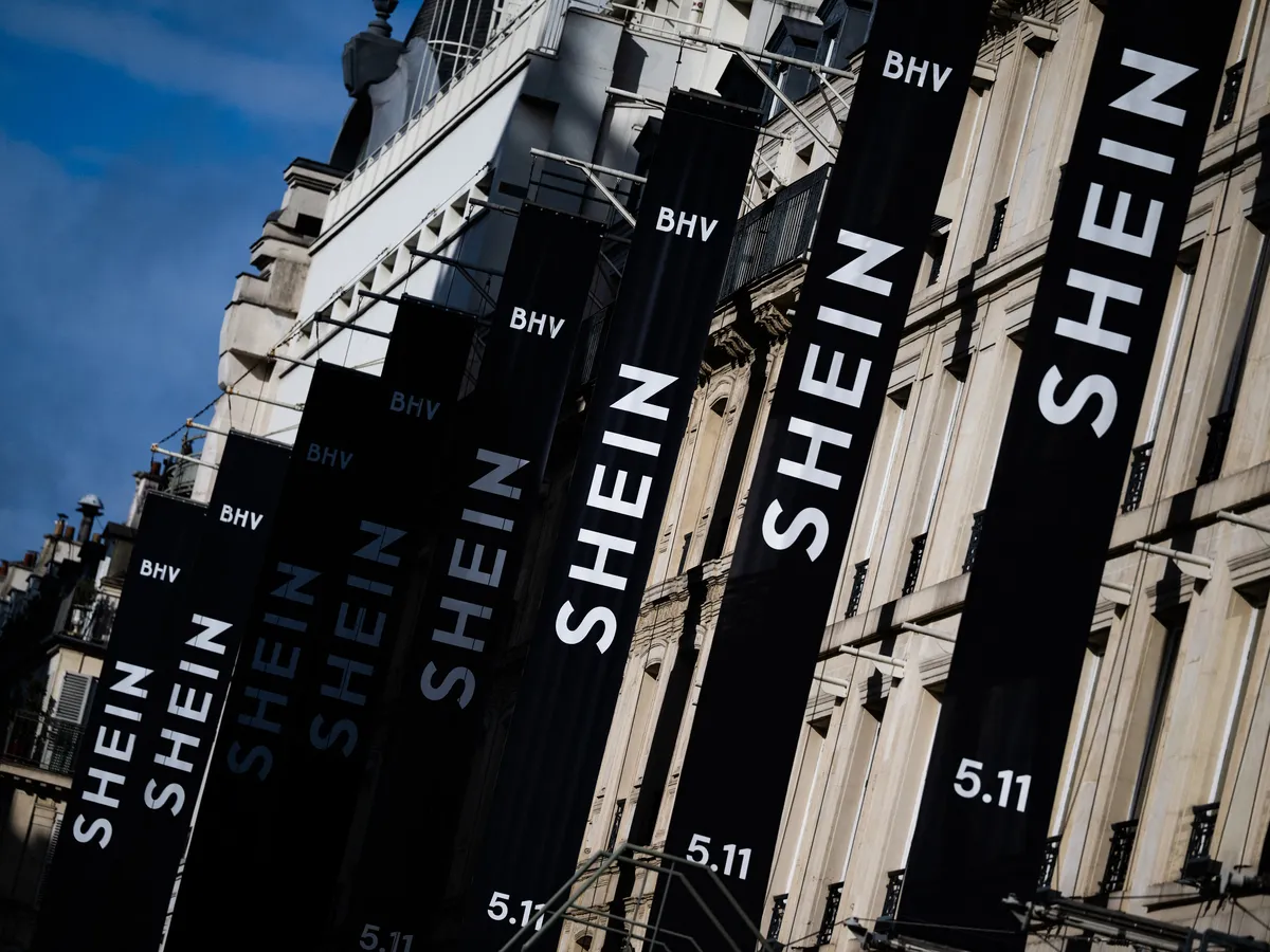 Tall black banners along a building spelling SHEIN with BHV logos and 5.11 at the bottom, against a blue sky.