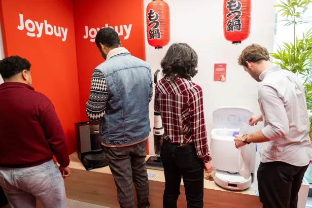 People stand at a red Joybuy display, examining kitchen gadgets and a white appliance on the right side of the scene.