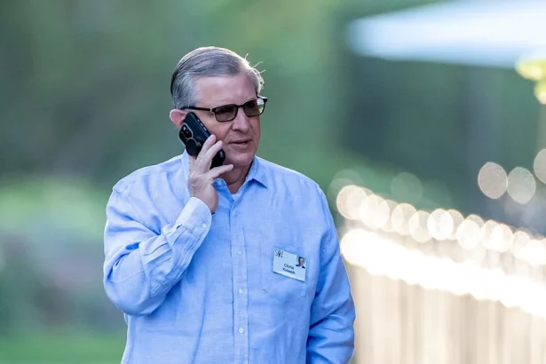 Older man with sunglasses and a blue button-up shirt on a mobile phone outdoors, with a name badge visible on his chest.