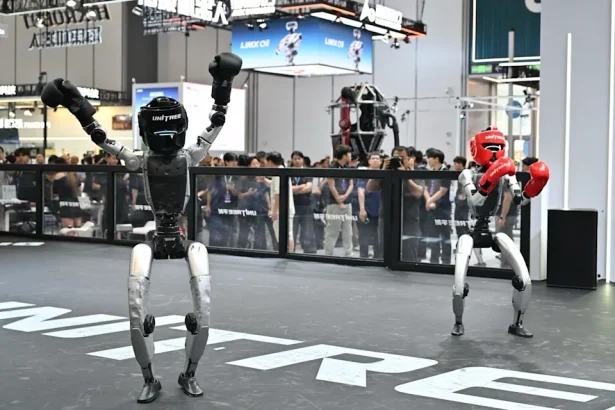Two humanoid robots boxing in a public arena, one with black gloves raising arms, the other with red gloves ready to strike, crowd watching behind a glass barrier.
