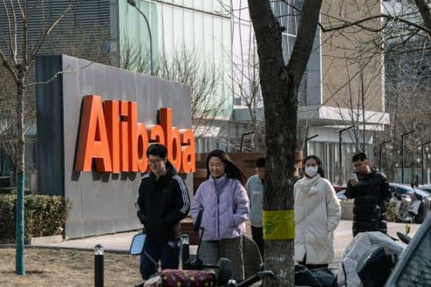 Group of people walk along a sidewalk near a large orange Alibaba sign on a corporate campus, with winter coats and some wearing masks.